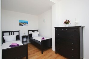 A room with two beds, a dresser, a small table, and a wall-mounted shelf with a plant.