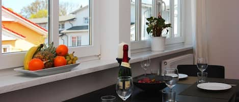 A dining area with a table set for two, a bottle of champagne, and a fruit bowl on the windowsill.