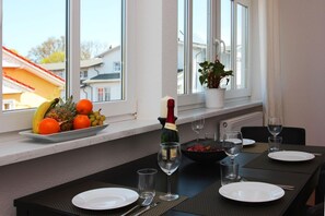 A dining area with a table set for two, a bottle of champagne, and a fruit bowl on the windowsill.