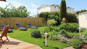 Um quintal com um deck de madeira, cadeiras de descanso e um jardim com diversas plantas e flores.
