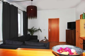 Una sala moderna con un sofá negro, un mueble de madera con un televisor y una mesa de comedor con un cuenco de fruta.