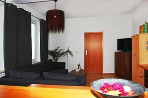 A modern living room with a black sofa, a wooden cabinet with a television, and a dining table with a bowl of fruit.