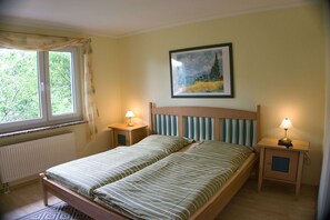 A bedroom with a double bed, two bedside tables with lamps, a window with curtains, a radiator, and a framed painting on the wall.