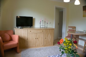 A living room with a TV on a wooden cabinet, a pink chair, a dining table with chairs, and a vase with flowers. - F-1042 Enna Vacation Apartment - Enna Vacation Apartment F-1042 Apartment 01 (Sellin)