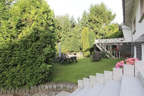Un jardin avec une table et des chaises pour les repas, entouré d’une végétation luxuriante et un escalier en pierre menant à une maison.