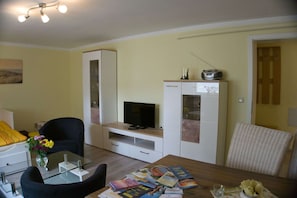 Uma sala de estar com televisão, uma mesa de jantar com revistas, uma cadeira preta, um armário branco e um travesseiro amarelo.