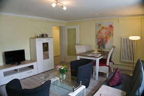 Una sala de estar moderna con televisor, muebles blancos, una mesa de centro de vidrio y un comedor con sillas y mesa.