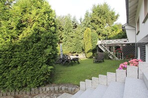 A backyard with a dining table and chairs, surrounded by lush greenery and a stone staircase leading to a house.
