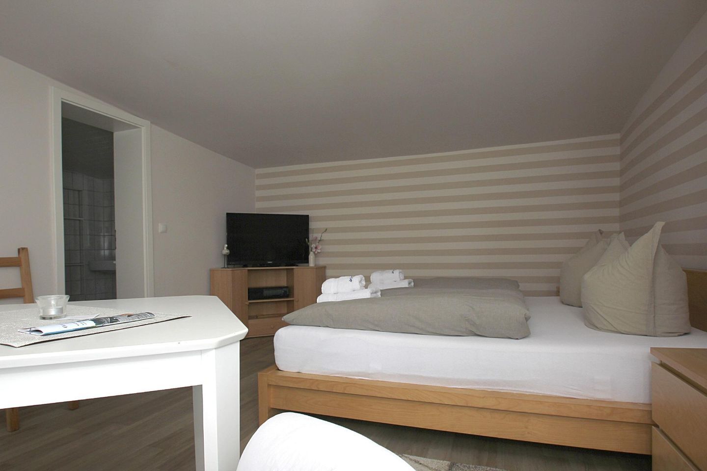 A hotel room with a bed, a TV on a wooden stand, a desk with a magazine, and a striped wall.