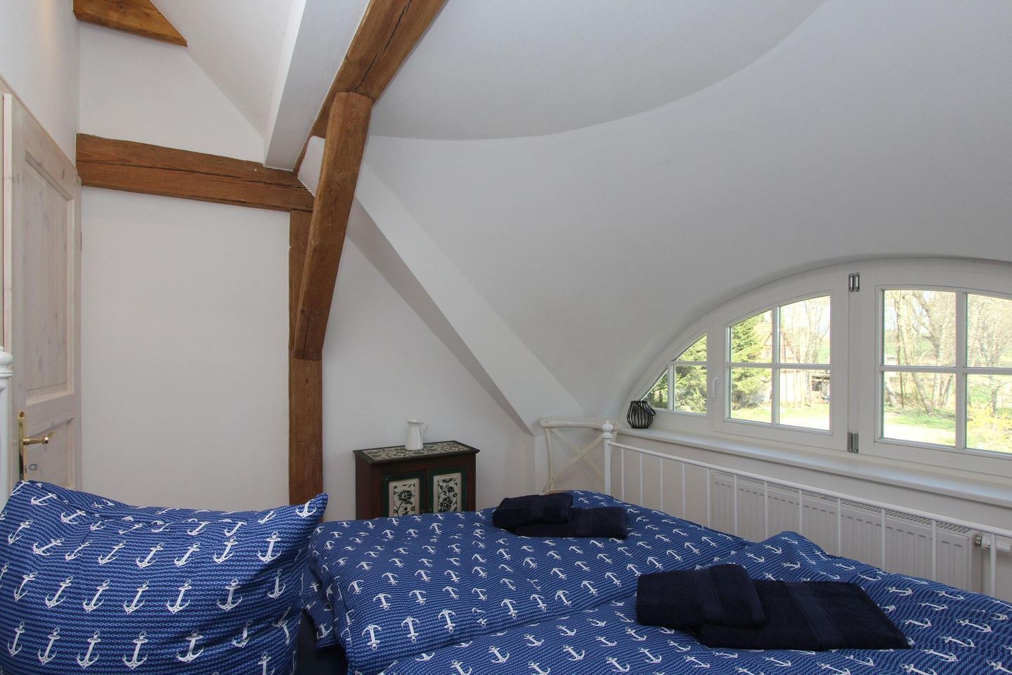 Ein Schlafzimmer mit schrägen Wänden, Holzbalken, einem Fenster mit Blick auf Bäume und einem Bett mit blauer Bettwäsche.