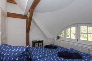 A bedroom with a sloped ceiling, wooden beams, a window with a view of trees, and a bed with blue bedding. - F-1081 Pfarrhof Vacation Apartment - Pfarrhof Vacation Apartment F-1081 (Garz/Rügen)