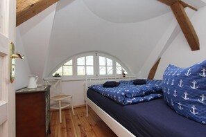 Un dormitorio pequeño y acogedor con techo inclinado, una cama individual con ropa de cama azul, una silla de madera y una mesita con una jarra.