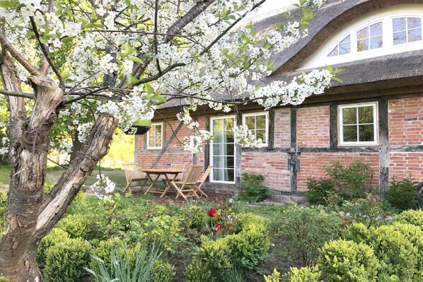 Une maison traditionnelle en briques, avec un toit de chaume, une table et des chaises en bois dans le jardin, et des arbres en fleurs.
