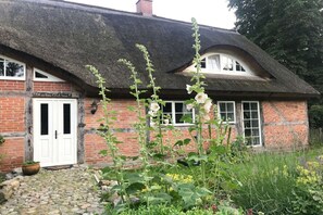 Une maison traditionnelle en briques, avec un toit de chaume, une porte blanche et un jardin où poussent diverses plantes et fleurs.