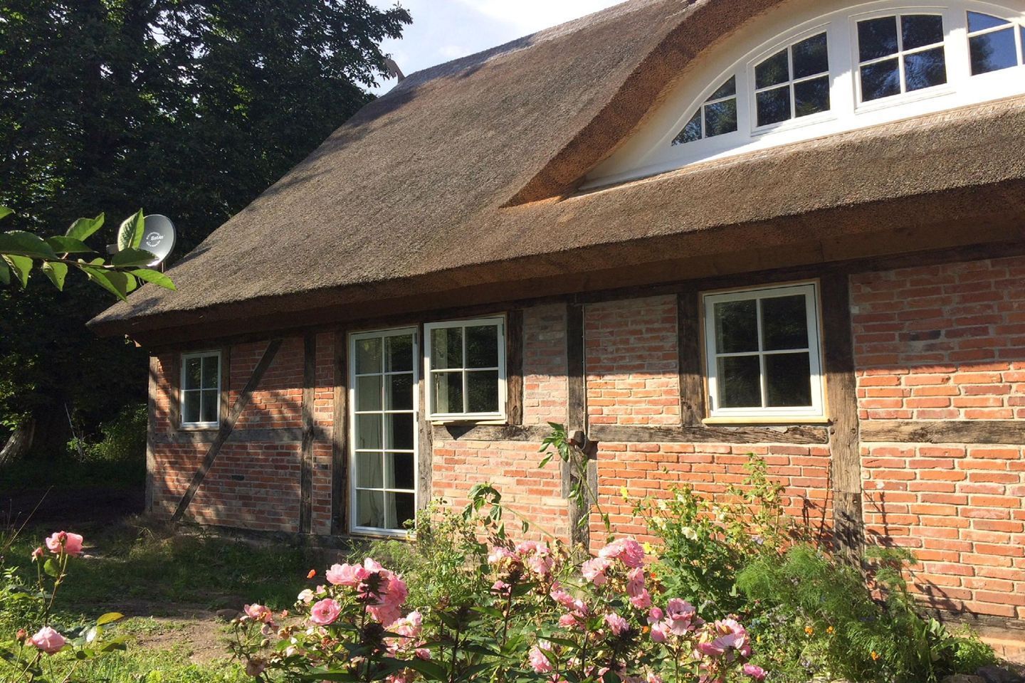 A traditional thatched-roof house with a brick exterior, multiple windows, and a garden with pink flowers.