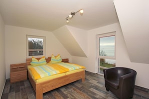 Ein Schlafzimmer mit schrägem Dach, einem Bett mit gestreifter Bettwäsche, einem Nachttisch, einem Stuhl und Meerblick durch ein Fenster.