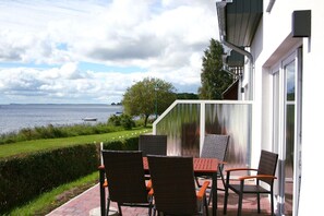 Eine Terrasse im Freien mit Tisch und Stühlen, mit Blick auf ein Gewässer und eine Rasenfläche.