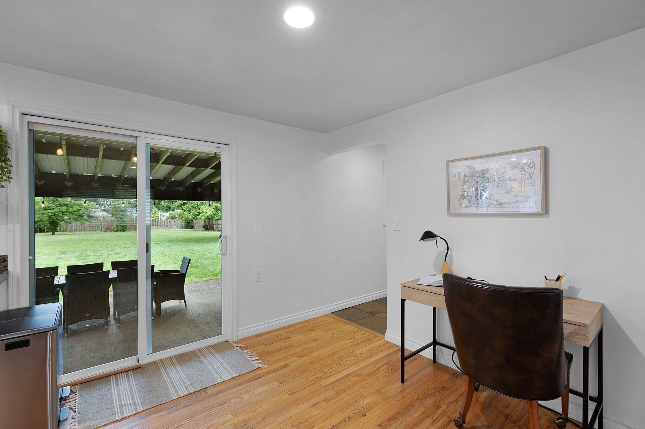Une pièce avec un plancher en bois, un bureau avec une chaise, une porte-fenêtre coulissante donnant sur un jardin et une carte encadrée accrochée au mur.