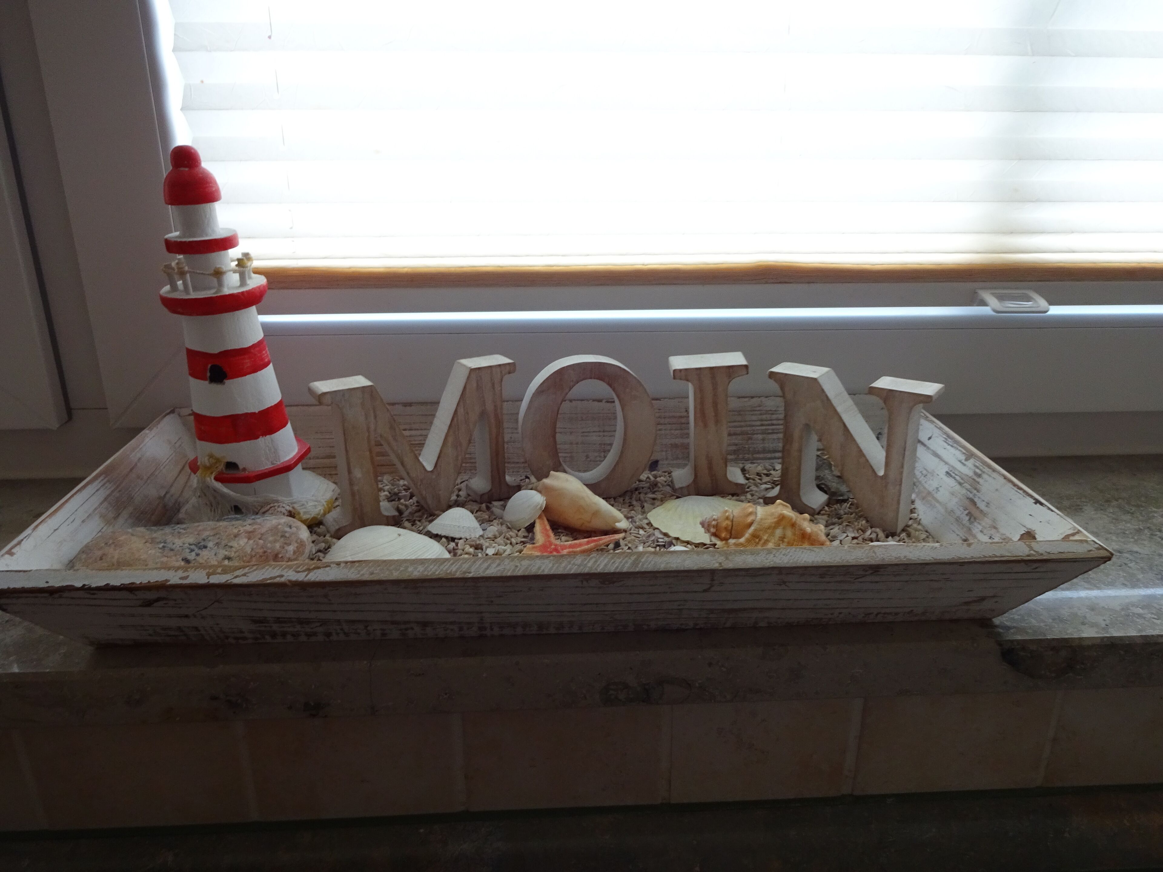 A decorative wooden sign with the word "MOIN" surrounded by seashells and a lighthouse model.
