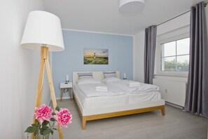 Une chambre à coucher comprenant un lit, des tables de chevet, une lampe, un tableau, une fenêtre avec des rideaux et un arrangement floral.