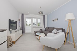 A modern living room with a grey sofa, a white armchair, a small round table, and a floor lamp. There is a television on a white cabinet, and a window with curtains.