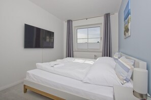 A neatly made bed with white linens, a mounted flat-screen TV on the wall, a window with curtains, and a bedside lamp. - Free use of the AHOI adventure pool and sauna - Haus Inselwind FeWo MEERbodden - (Gager)