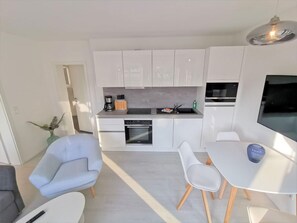 A modern kitchen with white cabinets, a built-in oven, and a microwave. There is a dining area with a white table and chairs, and a blue vase on the table. - Modern vacation apartment with a private terrace - Inselwind FeWo MEERhafen - (Gager)