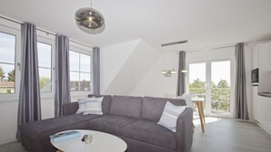A modern living room with a grey sectional sofa, a round white coffee table, and a large window with grey curtains. - With sea view, use of swimming pool and sauna - Haus Inselblick FeWo MEERrauschen (Gager)