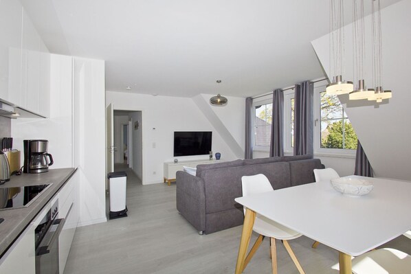 A modern living room with a grey sofa, a white dining table, and a TV in the background.