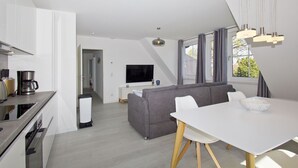 A modern living room with a grey sofa, a white dining table, and a TV in the background. - With sea view, use of swimming pool and sauna - Haus Inselblick FeWo MEERrauschen (Gager)