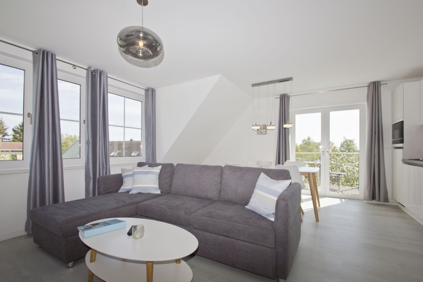 Un salon moderne avec un canapé d’angle gris, une table basse ronde blanche et une grande fenêtre agrémentée de rideaux gris.
