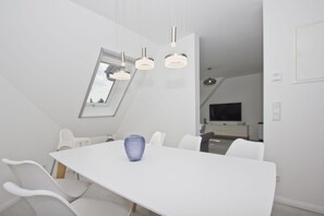 Een modern, wit interieur met een lange eettafel, witte stoelen en een blauwe vaas.