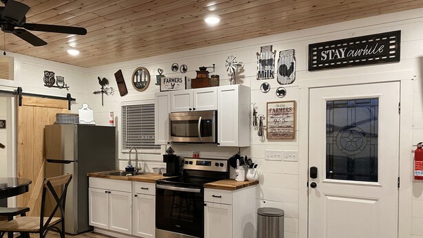 Una cocina moderna con armarios blancos, electrodomésticos de acero inoxidable y encimeras de madera. Hay un cubo de basura negro y una puerta con ventana.