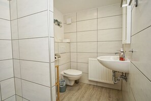 Un baño con paredes de azulejos blancos, inodoro blanco, lavamanos blanco y suelo de madera.