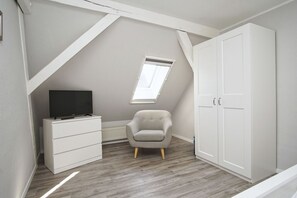 A room with a sloped ceiling, a white wardrobe, a grey armchair, a white dresser, and a television on the dresser.