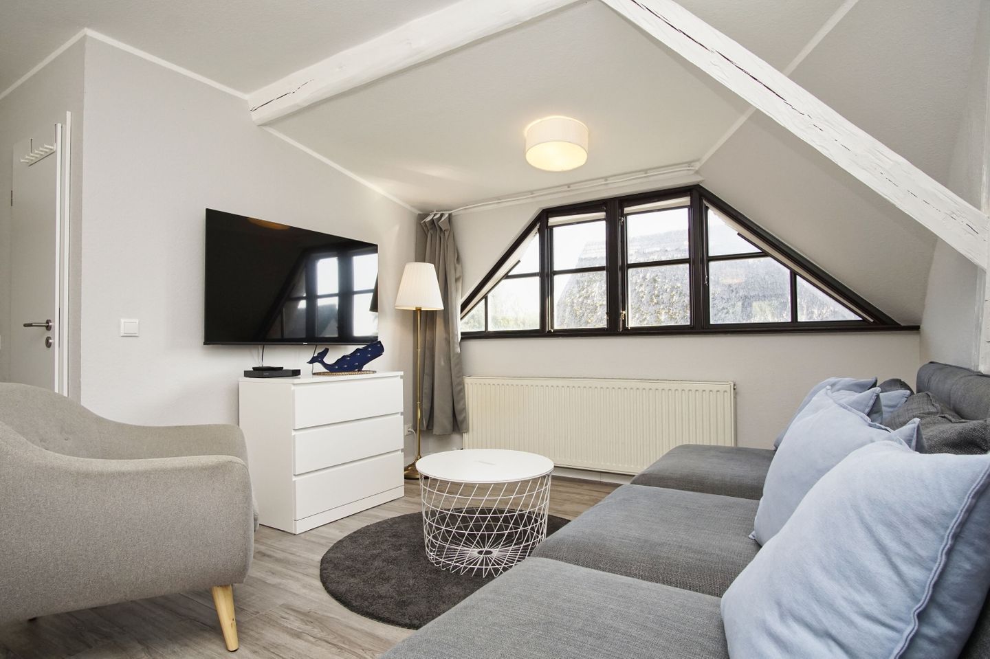 Quarto moderno estilo loft com teto inclinado, uma janela ampla, televisão de tela plana, cômoda branca, sofá cinza e mesa de centro redonda.
