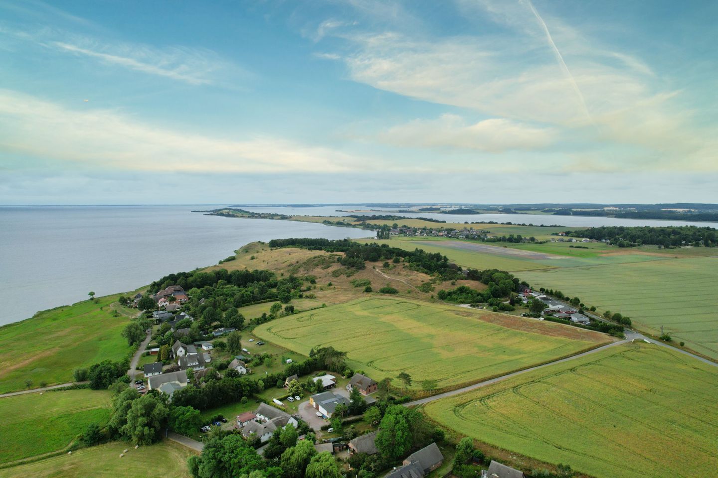 Oversigtsbillede af et landligt område med en lille landsby, grønne marker og et vandområde i baggrunden.