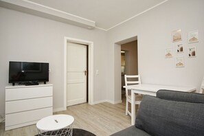 A compact living space with a flat-screen TV on a white cabinet, a grey sofa, a small round table, and a white dining table with a chair. - Close to the sea, quiet location, access to the pool and sauna - Haus Büdnerstube FeWo 2 (Middelhagen)