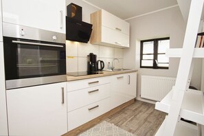A modern kitchen with white cabinets, a built-in oven, a coffee maker, and a window with blinds. - Close to the sea, quiet location, use of swimming pool and sauna - Haus Büdnerstube FeWo 2 (Middelhagen)