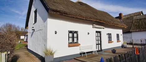 Een witte cottage met rieten dak, een houten bankje ervoor en een pad van bakstenen dat naar de ingang leidt.
