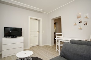 A compact living space with a flat-screen TV on a white cabinet, a grey sofa, a small round table, and a white dining table with a chair. - Close to the sea, quiet location, use of swimming pool and sauna - Haus Büdnerstube FeWo 2 (Middelhagen)
