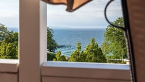 A view through a window of a coastal area with a pier extending into the sea, surrounded by lush greenery. - Fantastic sea view from the balcony, use of swimming pool and sauna - Villa Bergfrieden (Göhren)
