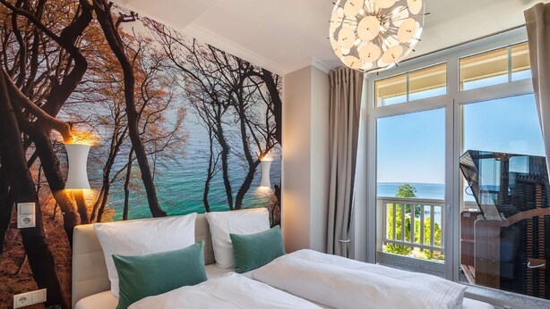 A bedroom with a large bed, two pillows, a nightstand with a book, a wall mural of trees and a sea view, and a balcony with a chair. - Fantastic sea view from the balcony, use of swimming pool and sauna - Villa Bergfrieden (Göhren)