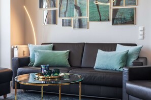 Uma sala de estar moderna com um sofá de couro escuro, almofadas verdes, uma mesa de centro de vidro com objetos decorativos e quadros nas paredes.