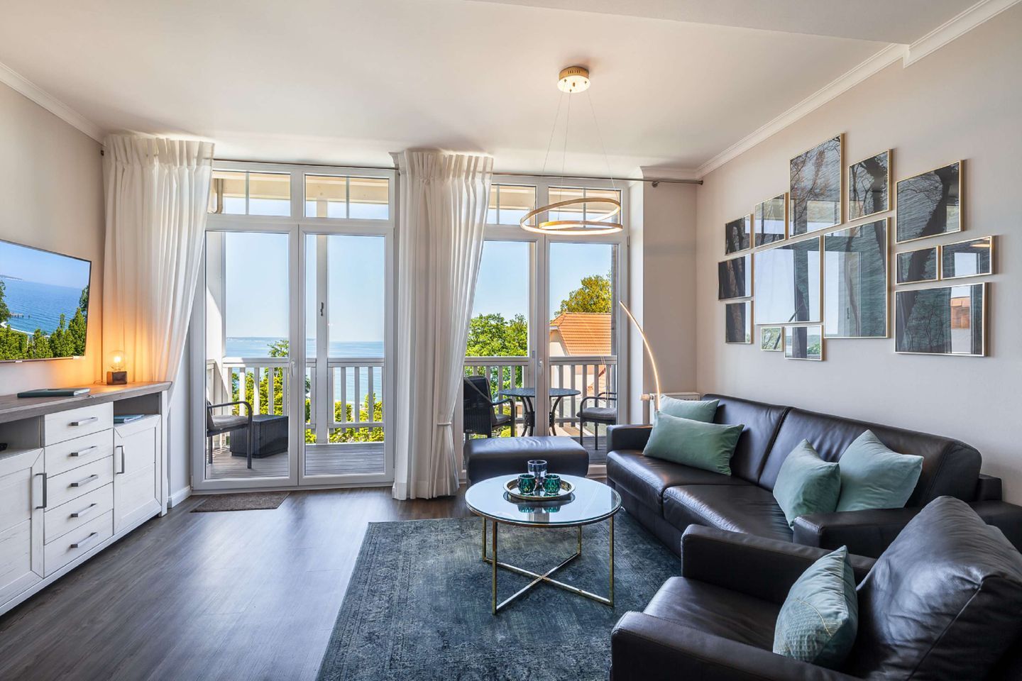 Un salon moderne avec un canapé, une table basse et un téléviseur. De grandes fenêtres sont agrémentées de rideaux, et il y a un balcon ainsi que des œuvres d’art accrochées au mur.
