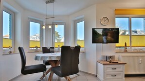 A modern kitchen with a dining area, large windows, a flat-screen TV, and a small cabinet with a radio.