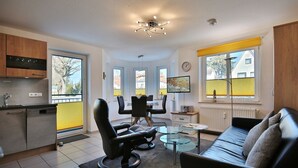 A modern kitchen with a dishwasher, dining area with chairs and a table, a sofa, and a TV on a stand.