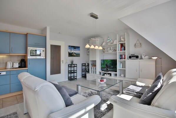 A modern living room with a glass coffee table, white sofas, a TV, and a kitchen area with blue cabinets.