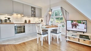A modern kitchen with white cabinetry, a dining area with a wooden table and white chairs, and a television displaying an elephant. - Am Elchgrund Apartment 12 - Im Brook 36-38 (Timmendorfer Strand)
