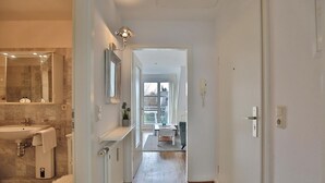 A modern bathroom with a marble sink, a mirror, and a shower. A hallway leads to a dining area with a table and chairs. - Am Elchgrund Apartment 12 - Im Brook 36-38 (Timmendorfer Strand)